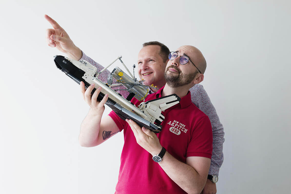 Un homme qui s'apprête à lancer un avion en Lego en l'air et un deuxième homme derrière lui qui regarde vers le haut en souriant